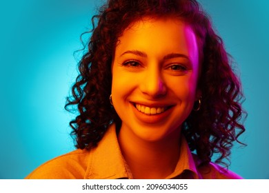 Cute Curly Model. Close-up Young Beautiful Cheerful Girl Smiling Isolated On Blue Studio Background In Neon Light. Concept Of Emotions, Facial Expression, Youth, Aspiration.