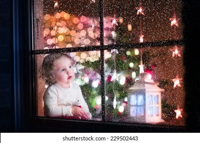 Cute Curly Toddler Girl Sitting With A Toy Bear At Home During Christmas Time, Preparing To Celebrate Xmas Eve, View Through A Window From Outside Into A Decorated Dining Room With Tree And Lights 