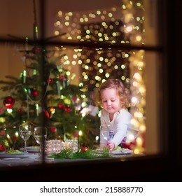 Cute Curly Toddler Girl Standing At A Christmas Dinner Table Settling The Dishes Preparing To Celebrate Xmas Eve, View Through A Window From Outside Into A Decorated Dining Room With Tree And Lights 