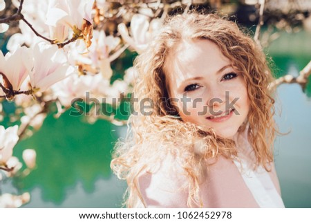 cute curly-haired girl posing in the park in the rays of the spring sun, enjoying magnolia and a pond