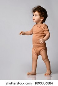 Cute Curly-haired Kid In A Beige Body Is Taking Its First Steps, With Ineteresmo Looking Up Against The Gray Background, Side View