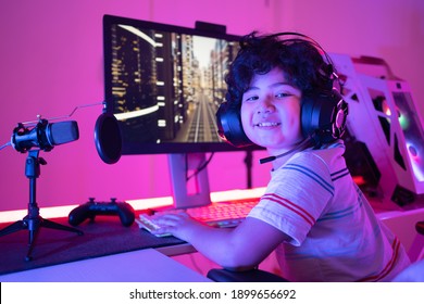 Cute Curly-haired Little Asian Boy Gamer In Headphones Sitting Playing Video Games On PC With RGB Keyboard Next To Joystick And Desktop Microphone Turning To Smile At The Camera In A Violet Neon Room.