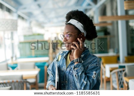 Cute dark skinned female entrepreneur making call while visiting coffee shop, inviting a candidate to a job interview.