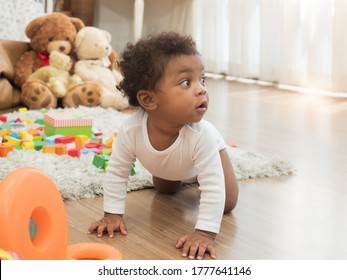 Cute dark skinned little African boy with curly hair crawling around the room, full of teddy bears and colorful toys, looking to his left side.