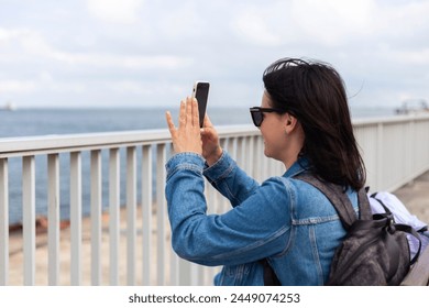 A cute dark-haired girl in a denim jacket takes pictures on her phone while standing on the sea coast - Powered by Shutterstock