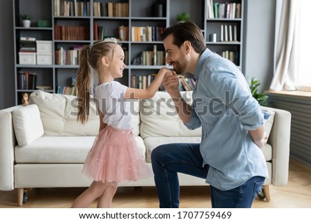 Cute daughter play with father indoor enjoy free time together, dad kneeling holds hand kisses arm of pretty kid girl showing good manners, politeness, gentlemen and little princess having fun at home