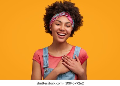 Cute delighted female with positive expression, keeps hands on heart as expresses her friendly manners, has positive smile, wears denim overalls and stylish headband, poses against yellow background