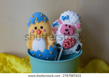 Cute Easter marshmallow lollipops featuring a yellow chick hatching from a blue egg and a white lamb holding a decorated egg, displayed in a mint green bucket.