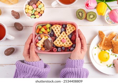 Cute Easter style lunchbox. Creative kids school lunch box with Easter symbols and sweets - bunny rabbit belgian waffle sandwiches, berry fruit, candy carrots, chocolate eggs - Powered by Shutterstock