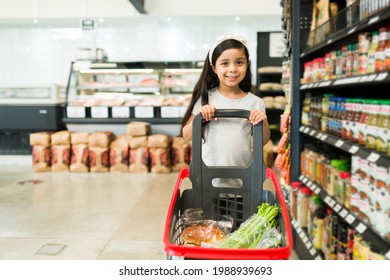 Cute elementary girl feeling happy while going grocery shopping alone at the supermarket and using a shopping cart