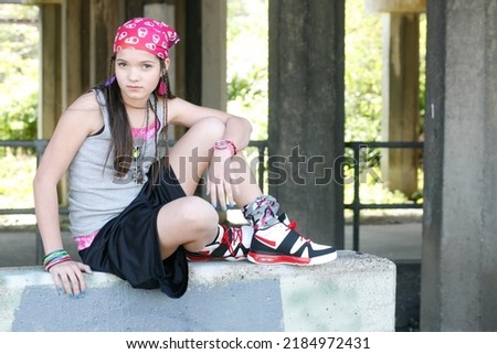 Cute ethnically ambiguous urban girl wearing a pink bandana, standing with arms crossed against a concrete wall. She looks like she could be a student, gang member, runaway, or juvenile delinquent.