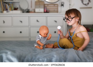 Cute European Girl Toddler Playing Doctor, Kid Wearing Toy Glasses With Stethoscope Listens To Doll, Child Treating Baby Doll