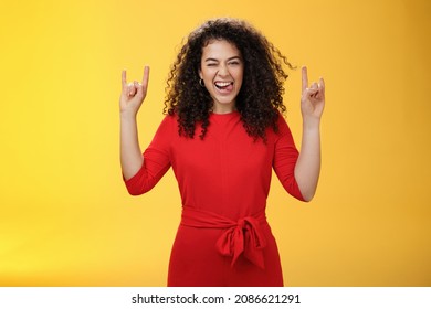Cute excied and daring woman in 25s sticking out tongue as fooling around having fun on concert winking joyfully and showing rock-n-roll gesture feeling amused and alive over yellow background