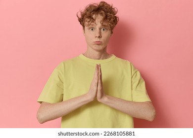 Cute excited begging curly haired ginger young man keeping hands clasped lips folded together with imploring look hopes for better says please dressed in yellow t-shirt isolated on pink background.