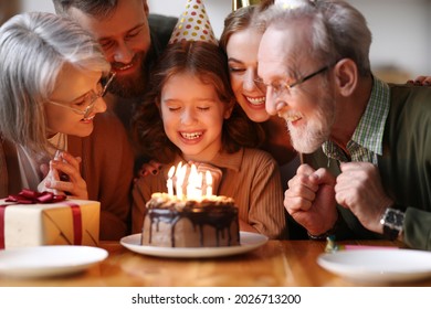 Cute excited little child making wish and blowing candles on cake while celebrating Birthday with big lovely family, positive parents and grandlarents in party hats congratulating happy girl at home - Powered by Shutterstock