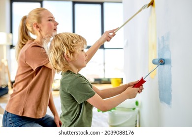 Cute Family Happy Mother Teaching Little Adorable Kid Boy How To Paint Wall Using Paintbrush Roller, Side View On Friendly Family In Casual Wear, Blue And Yellow Colours. Copy Space