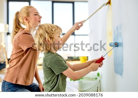 cute family happy mother teaching little adorable kid boy how to paint wall using paintbrush roller, side view on friendly family in casual wear, blue and yellow colours. copy space