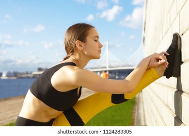 Cute Fashionable 20 Year Old Young Woman Choosing Active Healthy Lifestyle Doing Physical Exercise Outdoors, Stretching Leg Against Brick Walk. Sporty Pretty Girl Training By The River Alone