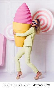 Cute fashionable woman  holding big props pink  ice cream, walking on hight heels  and laughing. Wearing bright elegant costume , stylish vintage  sunglasses.