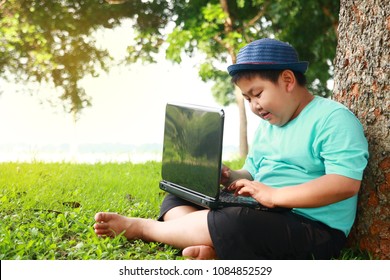 A cute, fat Asian boy wearing a blue shirt. Sitting to print a laptop computer under a big tree in the garden. He smiled happy. - Powered by Shutterstock