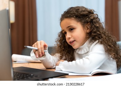 Cute Female Child Holding Pen And Watching Online Lesson On Laptop While Sitting At The Table At Home. Remote Education Concept 