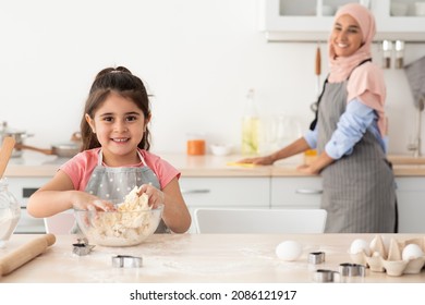 Cute Female Child Preparing Dough While Baking With Muslim Mom In Kitchen, Adorable Little Girl Learning How To Bake While Her Mother Cleaning Table On Background, Selective Focus On Kid