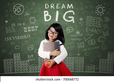 Cute Female Elementary School Student Standing In The Class While Holding A Book And Apple Fruit With Dream Big Text On Blackboard