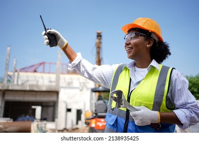 A cute female engineer of African descent, wearing a vest, a hard hat, holding a walkie-talkie, a wrench.A female worker with black skin is standing smiling.Behind her was a house under construction. - Powered by Shutterstock