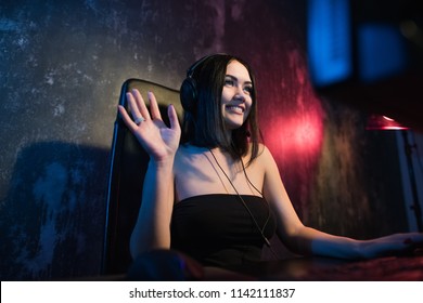 A cute female gamer girl sits in a cozy room behind a computer and plays games. Woman live streaming computer video games to her fans and shaking hand to them. Streamer and gamer concept
