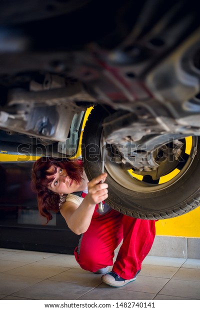 Cute, female mechanic in red work wear overalls\
in car mechanic shop fixing something under the car. Gender\
equality, diversity\
concept