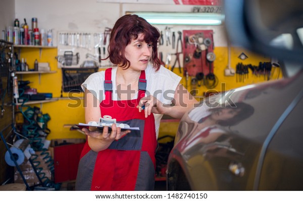Cute, female mechanic in red work wear\
overalls writes on clipboard in car garage. Car mechanic tools in\
the background. Gender equality, diversity\
concept