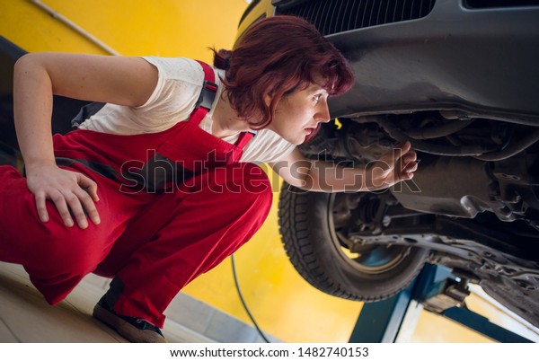 Cute, female mechanic in red work wear overalls\
in car mechanic shop looking under the car. Gender equality,\
diversity concept