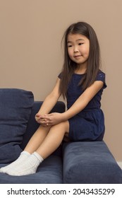 Cute Female Portrait Of Little Smiling Humble Girl, Pretty Adorable Asian Child Sitting On Blue Couch At Home, Vertical Image