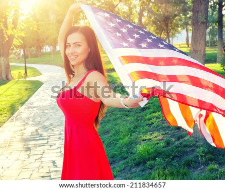 Cute female in red dress with American nation flag in her hands. Happy young woman holding american flag. Baclit shot at sunset