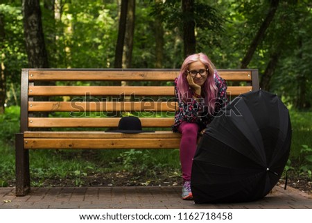 cute flirty smiling young girl with pink hair and glasses portrait on outdoor park environment sit on wooden bench with black classic hat and umbrella