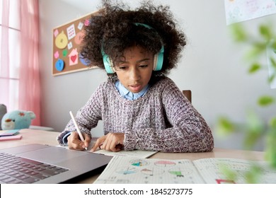 Cute focused african school kid girl wearing headphones virtual distance learning online listening remote education digital class doing homework studying at home classroom sitting at desk with laptop.