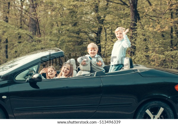 Cute four\
baby children friends of girls and boys people with at wheel\
pretends driving car as drivers on road\
outdoor