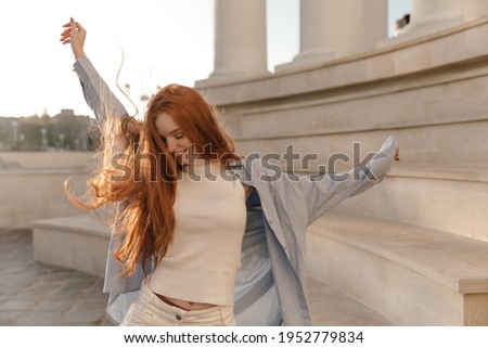 Cute foxy lady dancing outdoors at sunny day. Adorable young long-haired girl , wearing light turtleneck, blue shirt, moving and looking down against city landmark background