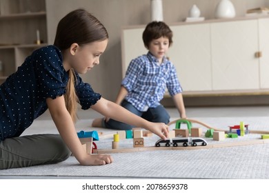 Cute Friendly Siblings Play Wooden Railroad Seated On Warm Floor In Living Room, Enjoy Playtime Together And Modern Plaything, Riding Toy Train On Rail Road. Hobby And Leisure At Home, Games Concept