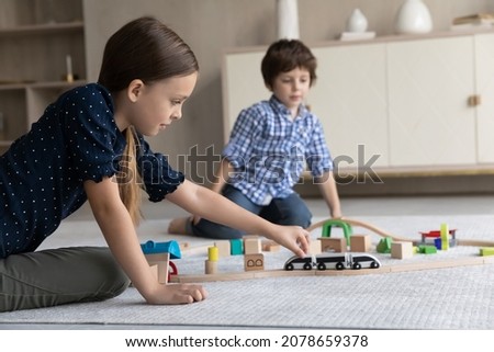 Cute friendly siblings play wooden railroad seated on warm floor in living room, enjoy playtime together and modern plaything, riding toy train on rail road. Hobby and leisure at home, games concept