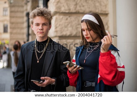 Cute fun curly teenage boy and girl wearing stylish 90s fashion clothes with chains around neck. Youth subculture. Happy teenage boy and girl wears stylish clothes with chains with phone for a walk.