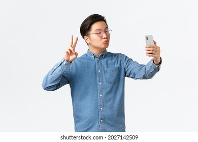 Cute And Funny Asian Young Guy Pouting Silly, Taking Selfie On Smartphone, Using Photo Filter App To Change Appearance, Shooting Himself With Peace Sign And Kiss, White Background