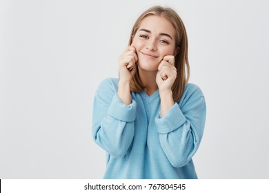 Cute funny attractive girl with straight fair hair pinching her cheeks and smiling, wearing blue sweater, posing against gray background. People and positive emotions concept.