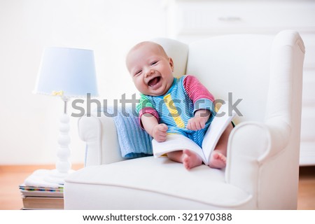 Cute funny baby boy reading a book sitting in a white chair at home. Children read books in a library seat. Nursery and playroom interior for kids. Early development and learning for young kid.