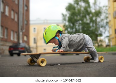 Cute Funny Baby In Green Helmet And Grey Clothes Lying On Belly On Skateboard And Pushing Himself By One Leg. Closeup Photo Of Little Skateboarder. Outside On Asphalt Street