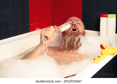 Cute funny bearded man taking bubble bath with rubber ducks and singing into bath brush enjoying relaxing at home after hard day.