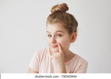 Cute Funny Blond Little Girl With Bun Hairdo Holding Finger Near Lips, Looking In Camera With Raised Eyebrows And Sneaky Face Expression.