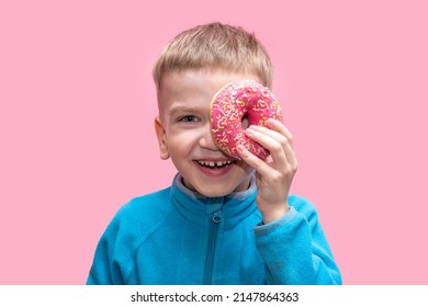 A cute funny boy in a blue sweater holds a bright pink donut near his eye and laughs on a pink background. Adorable cheerful kid is playing with a donut. Cheerful childhood concept.