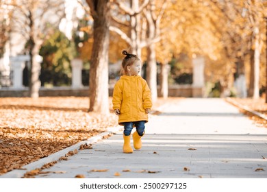 Cute funny child girl 3-4 year old wearing yellow raincoat and rubber boots walking over fallen leaves outdoor. Autumn season. Childhood. - Powered by Shutterstock