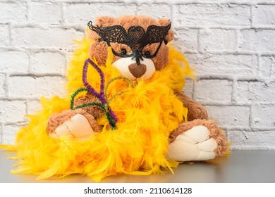 Cute funny fluffy brown teddy bear kids favourite stuffed toy with black carinaval mask and yellow feather boa sitting on shelf. Mardi Gras Party concept